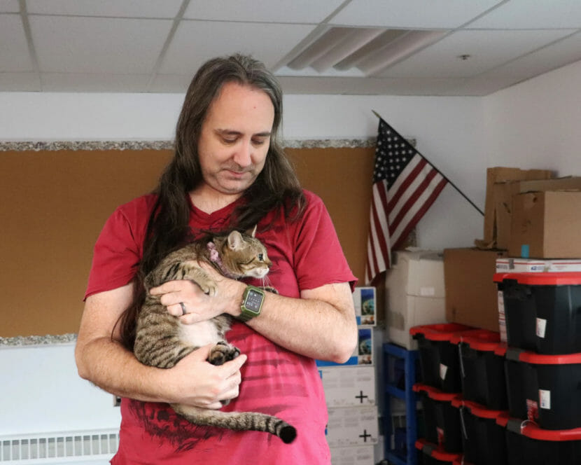 A man stands holding a cat in a classroom, with a large stack of totes nearby