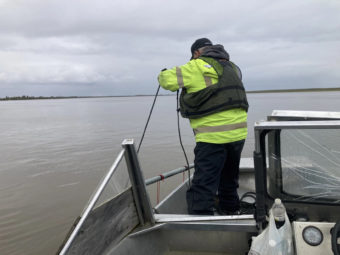A man stands in the bow of a boat in a wide river, dangling something into the water