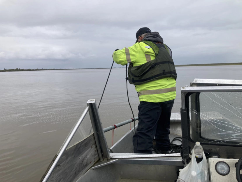 A man stands in the bow of a boat in a wide river, dangling something into the water