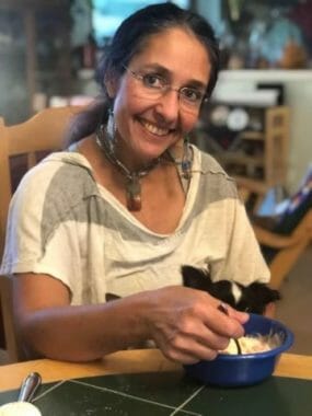 A smiling woman sits at a table holding a spoon in a bowl of something you can't see