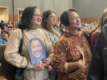 Women standing in an audience, looking toward the stage. One his holding a cutout photo of Mary Peltola's face