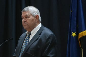 Charlie Pierce, wearing a suit, stands at a lectern next to an Alaska flag