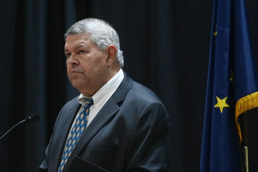 Charlie Pierce, wearing a suit, stands at a lectern next to an Alaska flag