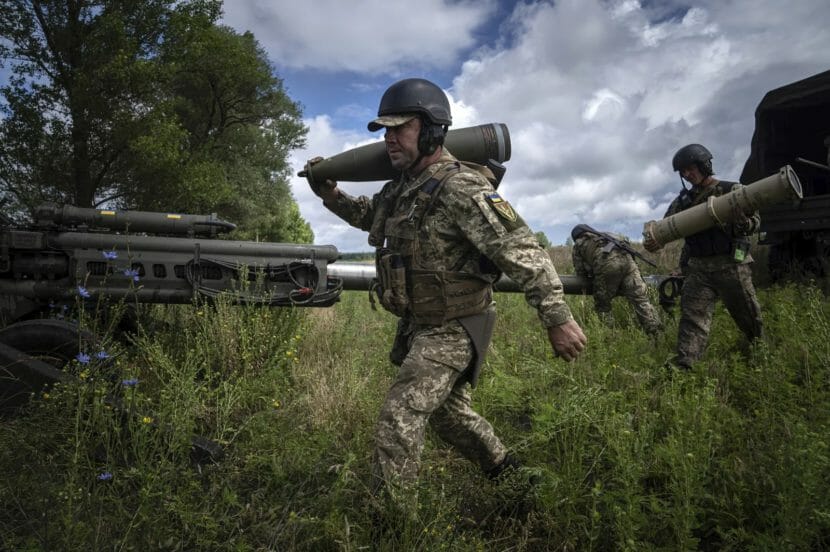 Three men in camouflage working around a field gun, one of them carrying a large artillery shell