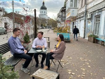 Three young people sit talking at an outdoor coffee shop table.