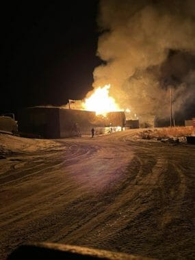 A man walks along the side of a building that is engulfed in flames.