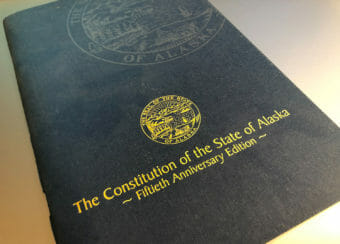 A blue copy of the Alaska Constitution booklet with yellow writing.
