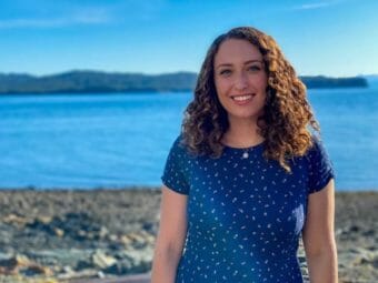 A portrait of a young woman standing on a beach with the water behind her.