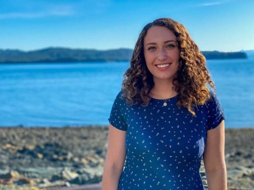 A portrait of a young woman standing on a beach with the water behind her.