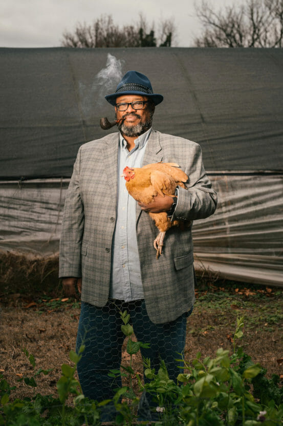 A man in a fedora smokes a pile while holding a live chicken.