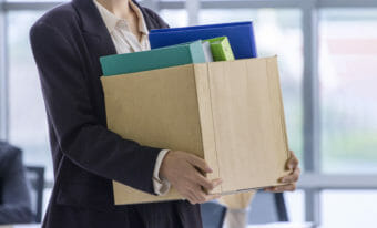 A stock photo of a person in a suit leaving an office carrying a cardboard box full of binders.
