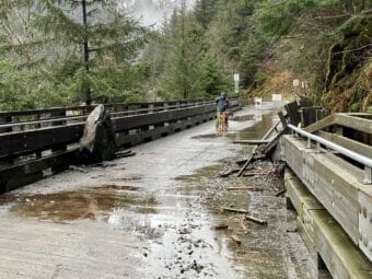 A man walks across an old trestle with a damaged railing and a boulder lying on it.