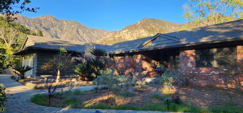 A ranch home made of brick sits among trees with mountains behind