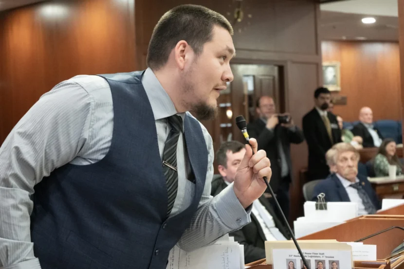 A man speaks into a microphone in a legislative chamber. 