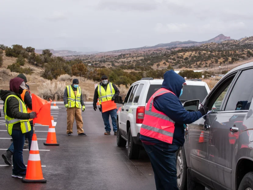 People i high-vis vests and masks speak to people waiting in cars. 