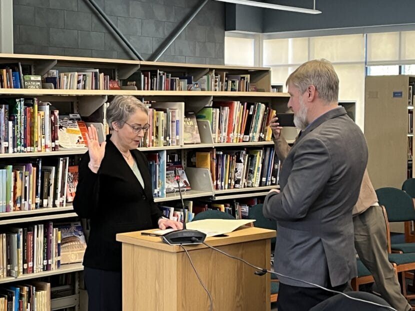 A woman dressed in black stands over a wooden podium with her right hand raised. A man in a gray suit stands across from her.
