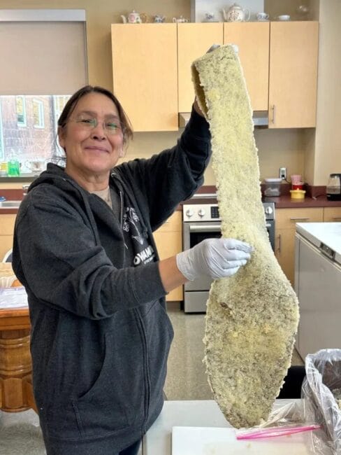 A woman standing in a kitchen smiles while a long strand of seaweed covered in herring eggs