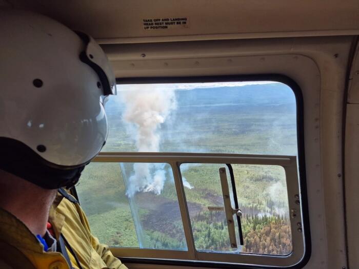 A man in a flight helmet looking through an aircraft window at a wildfire below.