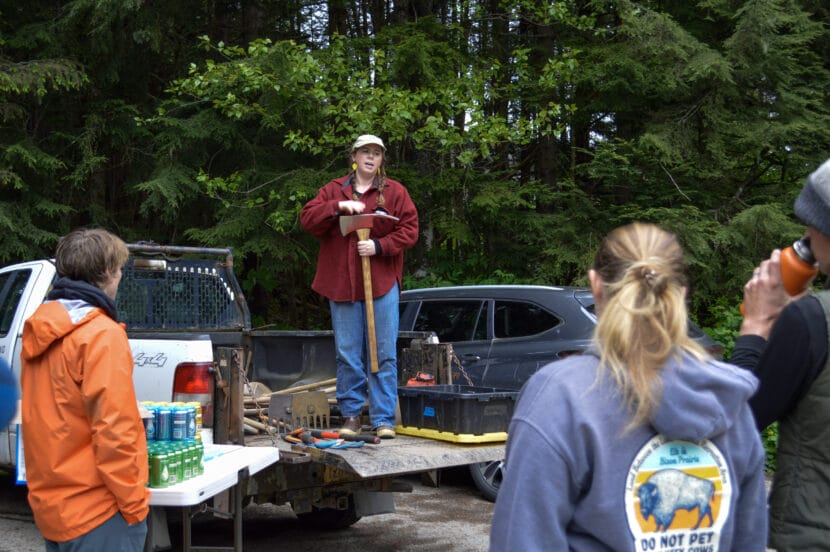 Trail Mix Inc Director Meghan Tabacek holds a Pulaski as she demonstrates safe tool usage to volunteers. She's standing on the bed of a pickup truck, wearing a red halibut jacket and lemon earrings.