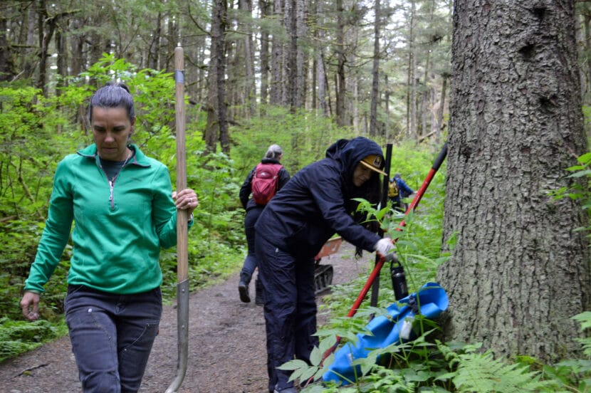 Two women Ami Reifenstein and Maggie McMillan hold tools on a trail on a forested path.