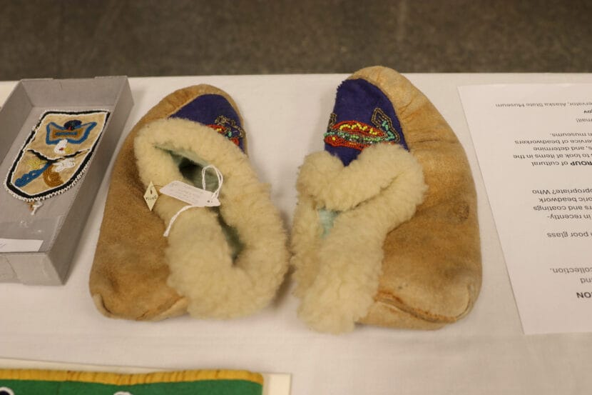 A pair of tan moccasins are laid on a white table. The top of the moccasins are dark blue with multicolored beadwork.