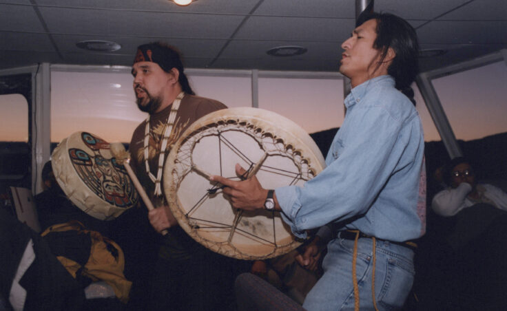 Two men drum for an audience inside a whale-watching boat. From the Cyril George Photo Collection.