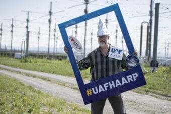 A man wearing a tall, pointed tinfoil hat stands in front of a field of giant antennas, holding an oversized picture frame that says hashtag UAFHAARP on it.