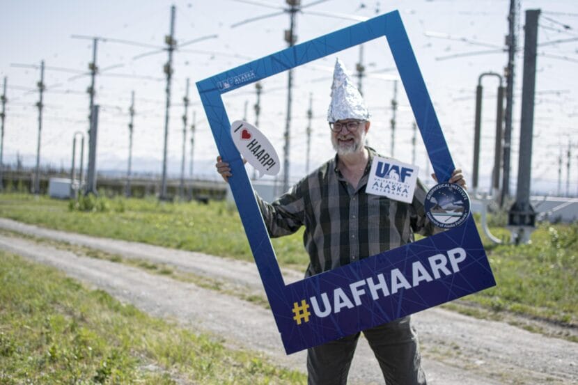 A man wearing a tall, pointed tinfoil hat stands in front of a field of giant antennas, holding an oversized picture frame that says hashtag UAFHAARP on it.