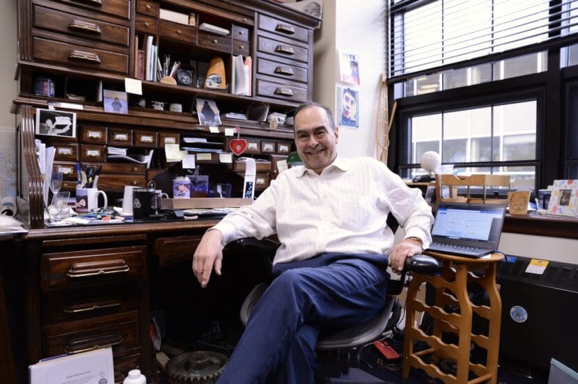 A man in a white dress shirt and blue pants sits in a chair in an office