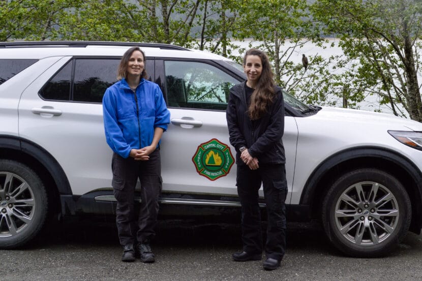 Two women stand smiling by a car marked "Capital City Fire/Rescue." Meghan DeSloover and Sarah Zaglifa respond to mental health emergencies in Juneau as part of a new mobile crisis team. July 1, 2025. (Photo by Yvonne Krumrey/KTOO)
