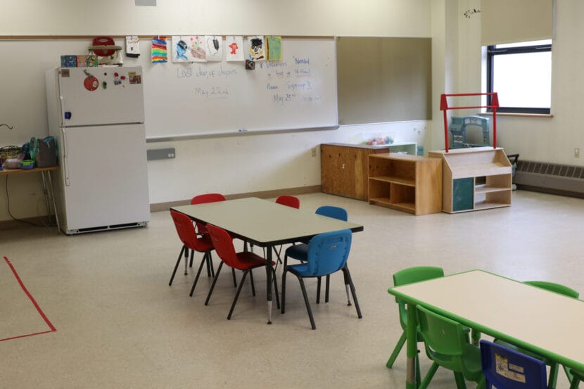 Red, blue and green chairs around small off-white tables in an empty classroom.