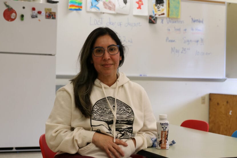 A woman in a white sweatshirt and blue glasses sits in a classroom.
