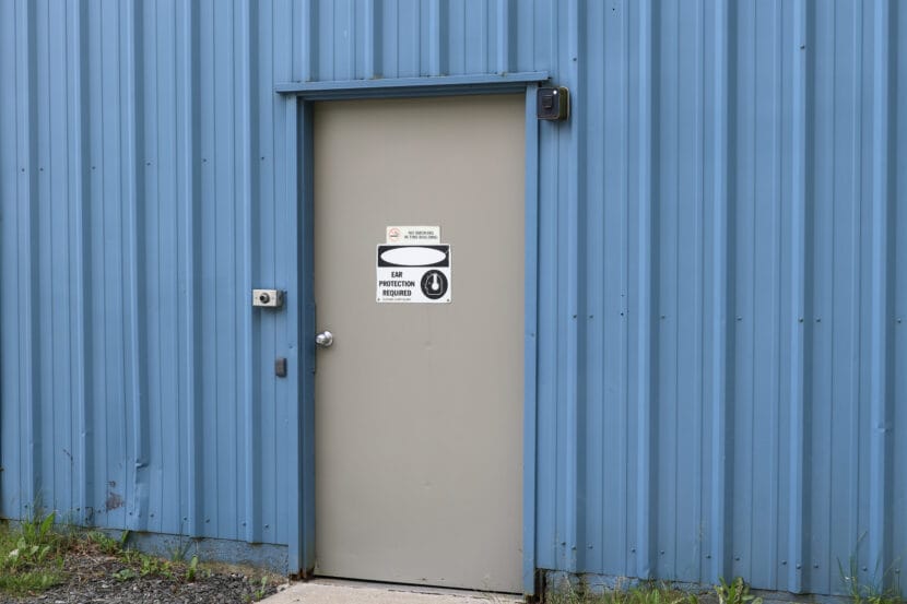 A gray door against a blue metal building with the sign "Ear Protection Required."
