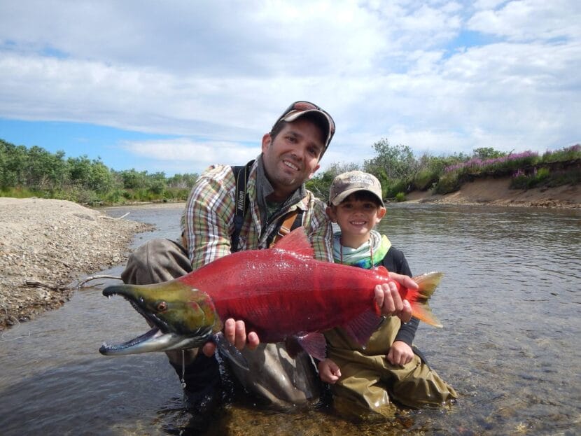 Donald Trump Jr. and his son in river shallows. Trump jr. holds in front of him a sockeye salmon that is bright red with a green head.