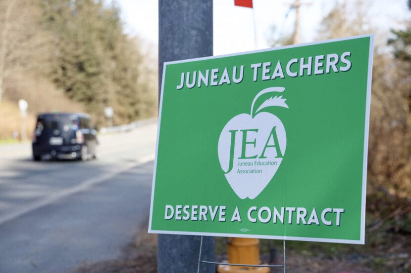 A green and white lawn sign with the Juneau Education Association logo and text saying, "Juneau teachers deserve a contract" as a black car drives in the background.
