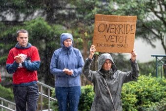 Suzanne Cohen holds a sign in the rain outside the Alaska Stae Capitol on Friday, Aug. 1 calling on lawmakers to override Gov. Mike Dunleavy's veto of more than $50 million in public school funding.
