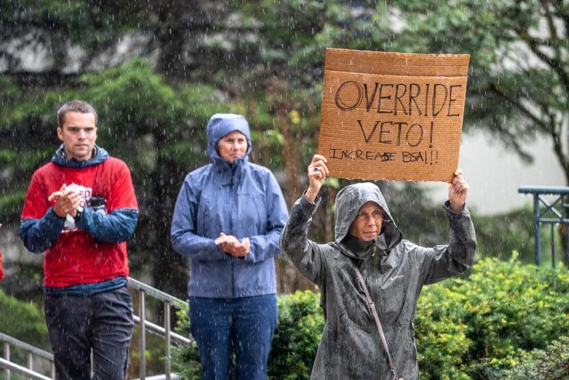 Suzanne Cohen holds a sign in the rain outside the Alaska Stae Capitol on Friday, Aug. 1 calling on lawmakers to override Gov. Mike Dunleavy's veto of more than $50 million in public school funding.