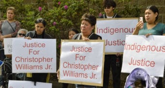 Five people hold signs that say "justice for Christopher Williams Jr." and "Indigenous Justice" in front of the brick exterior at the Juneau police station. Two wear button vests.