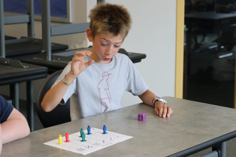 A child in a gray shirt looks at a purple dice with a raised hand. Small, multicolored game pieces are placed on a piece of paper to the side.