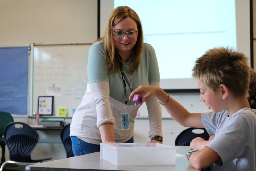 A woman leans on a gray desk and watches a child roll a large, purple dice.