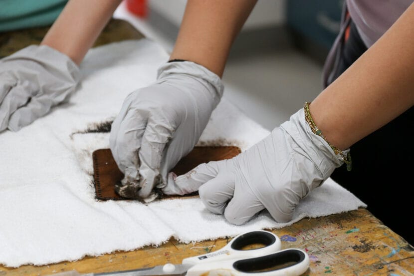 Gloved hands wipe dark brown liquid on a piece of leather on top of an off-white towel.