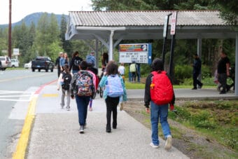 Several students walk on a sidewalk toward a covered walkway while carrying backpacks.