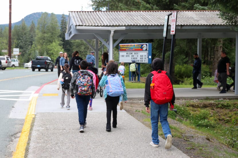 Several students walk on a sidewalk toward a covered walkway while carrying backpacks.