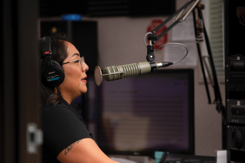A woman wearing headphones speaks into a radio studio microphone