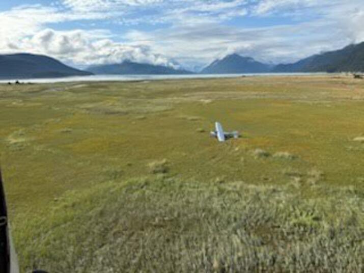 A remote coastal plain with mountains in the background. In the center of the image, a small airplane sits tilted leaning on its left wingtip.