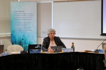 UA President Pat Pitney sits at a table lined with black cloth with a live microphone and laptop in front of her.