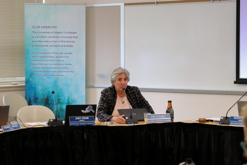 UA President Pat Pitney sits at a table lined with black cloth with a live microphone and laptop in front of her.