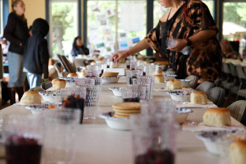 A student in a brown shirt sets clear plastic cups on a table full of individual servings of fish soup, berries and bread.
