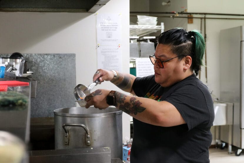 A teacher with green and black hair scoops soup from a large stock pot into a paper bowl.
