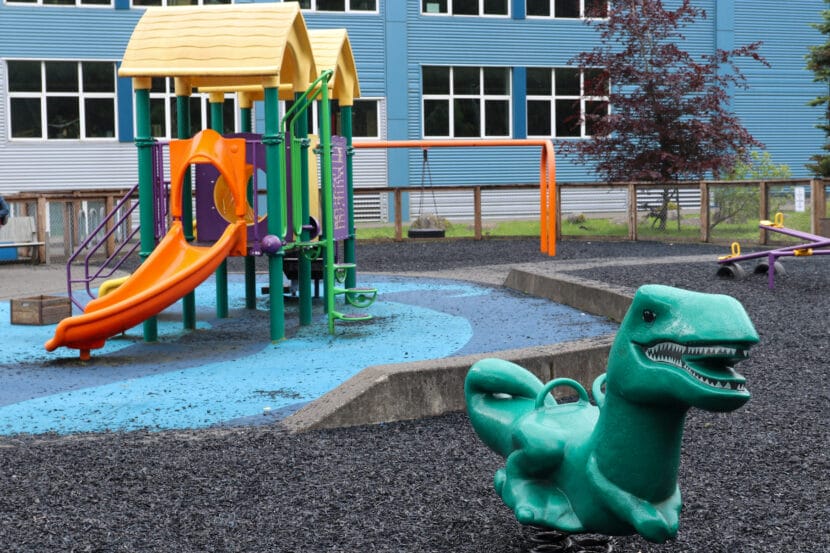 A green T-Rex on springs is on black shaved tires with a green and orange play structure in the background.